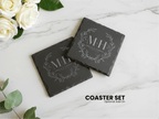 Two black slate coasters with engraved initials on a marble surface with white roses and green leaves.