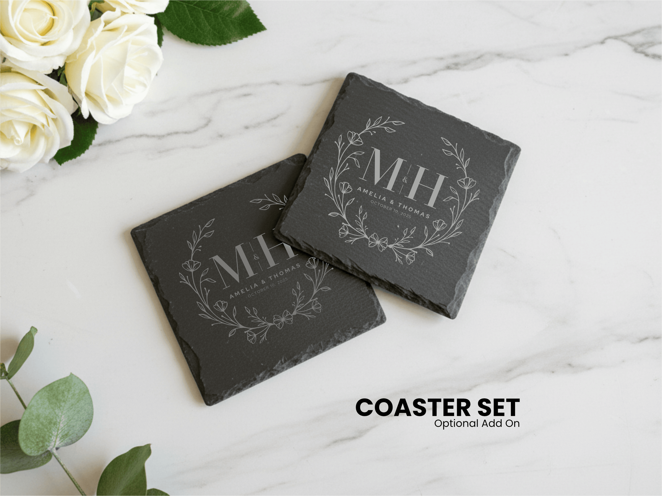 Two black slate coasters with engraved initials on a marble surface with white roses and green leaves.