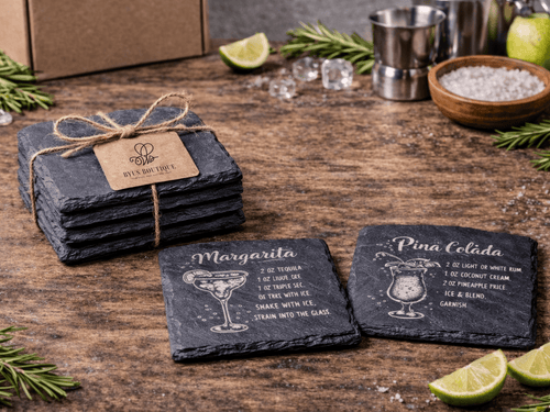 Set of slate coasters with cocktail recipes on a wooden surface with limes and a cocktail shaker.