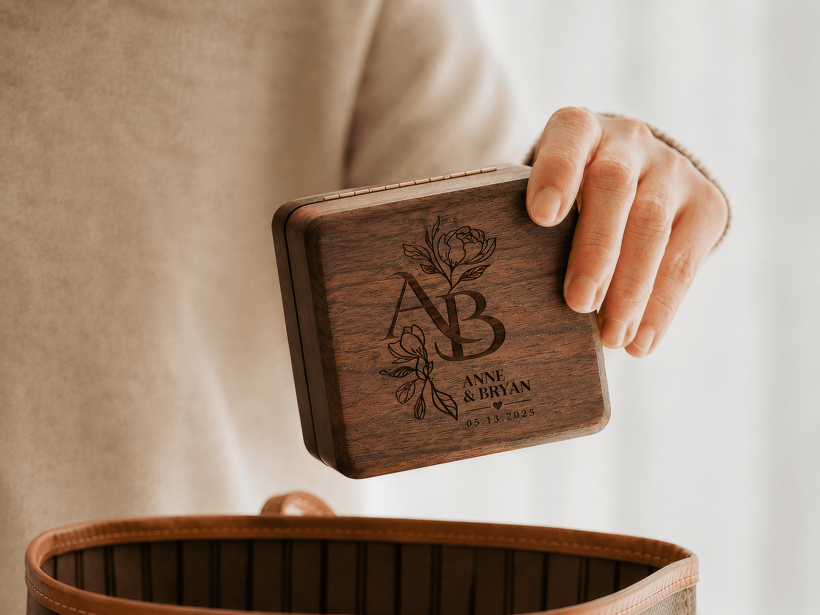 A hand holding a square dark wood keepsake box engraved with floral artwork and names over a beige sweater and a brown leather bag.
