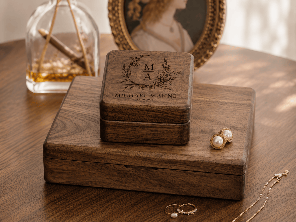 A personalized wooden jewelry box with engraved initials and a name sits on a larger wooden box on a wooden table. Gold jewelry, including pearl stud earrings and rings, is placed nearby, with a softly blurred vintage portrait and glass bottle in the background.