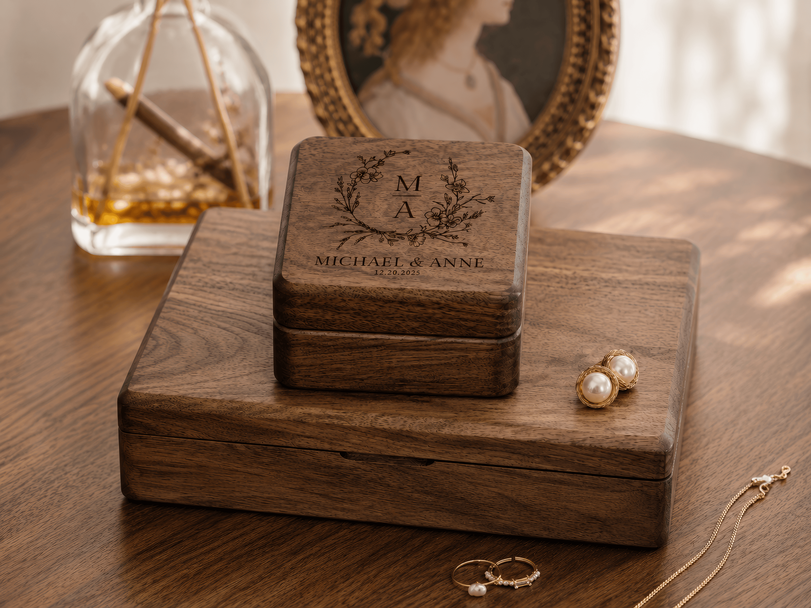 A personalized wooden jewelry box with engraved initials and a name sits on a larger wooden box on a wooden table. Gold jewelry, including pearl stud earrings and rings, is placed nearby, with a softly blurred vintage portrait and glass bottle in the background.