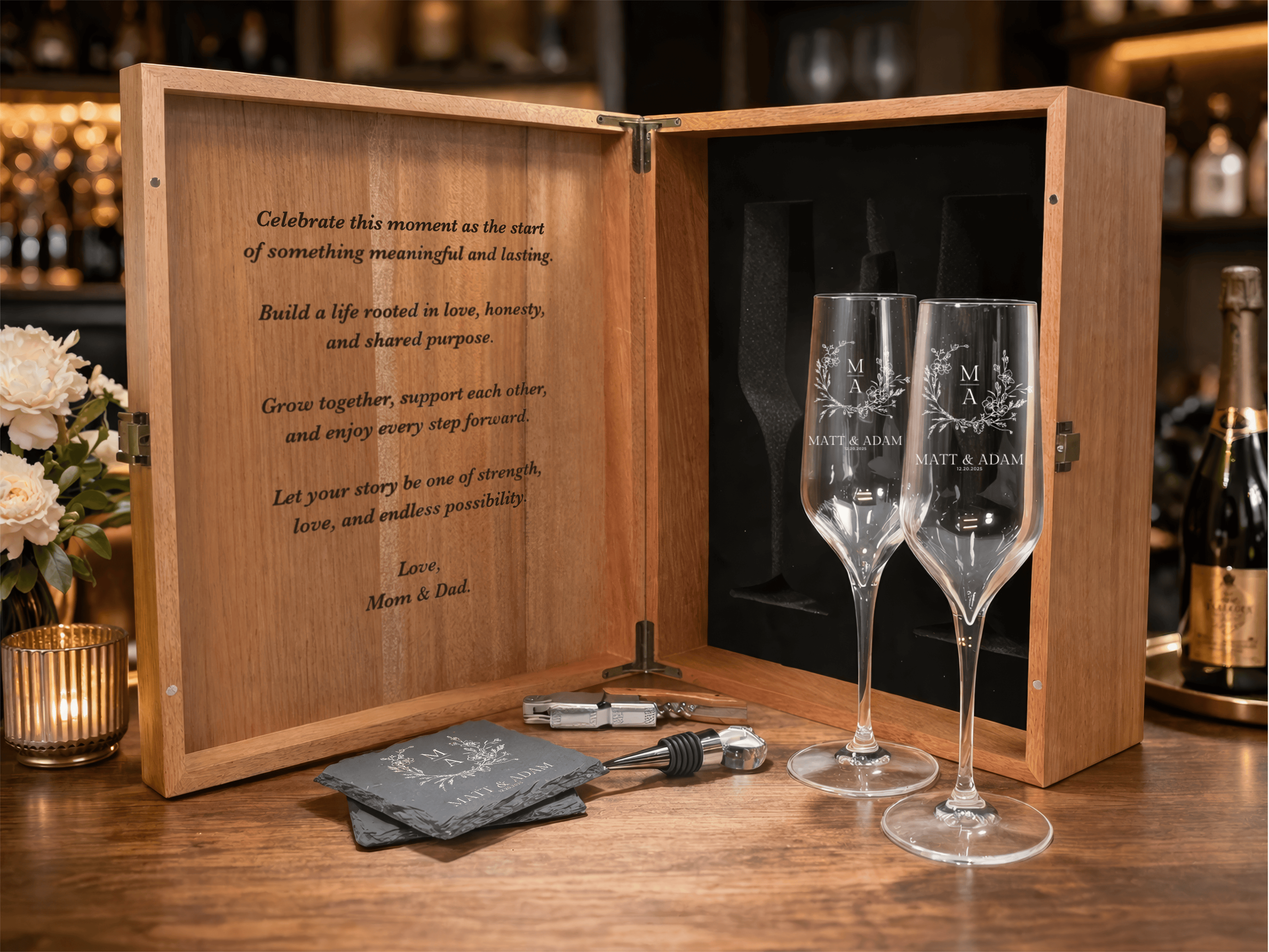 Two engraved wine glasses in a wooden box with a romantic message, surrounded by flowers and a bottle of champagne.