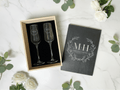 Two engraved champagne flutes in a wooden box with a matching cover on a white surface with flowers.