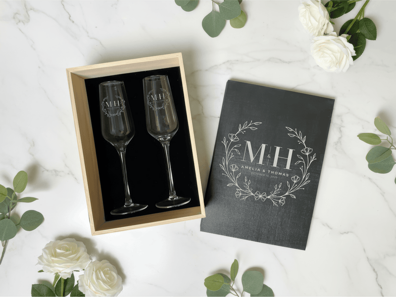 Two engraved champagne flutes in a wooden box with a matching cover on a white surface with flowers.
