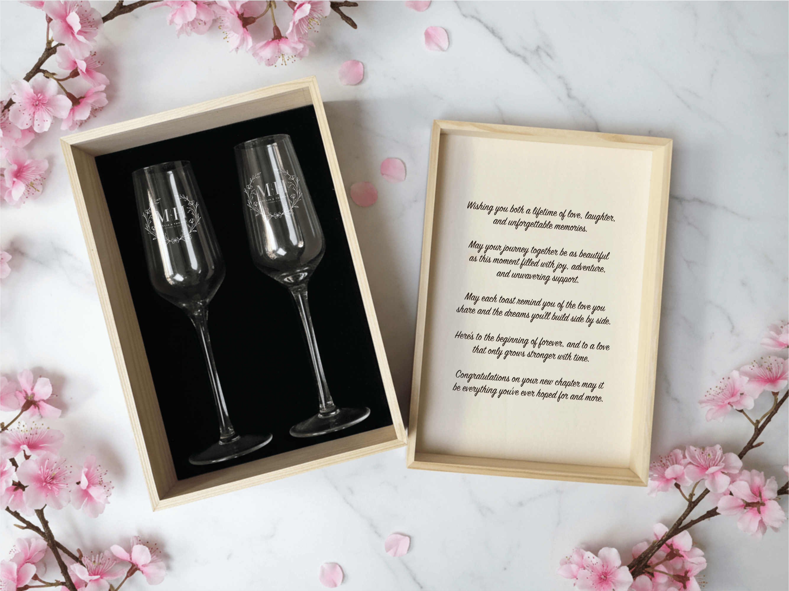 Two champagne glasses in a box with a framed poem on a marble surface with cherry blossoms.