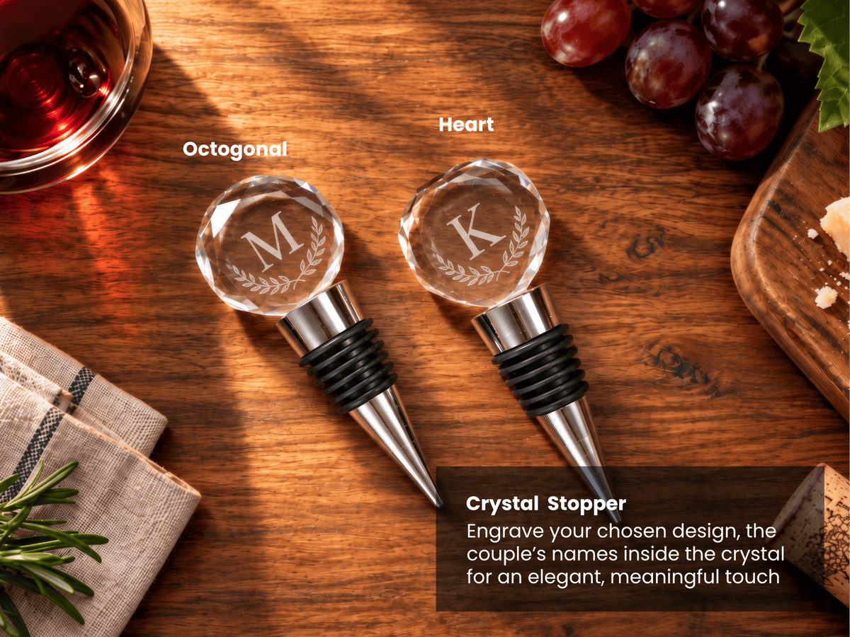 Two personalized crystal wine stoppers with engraved monogram letters 'M' and 'K' on faceted round tops, shown on a wooden tabletop with grapes, a glass of red wine, and a cutting board. Text labels identify the shapes as Octagonal and Heart, with a product description box at the bottom right.