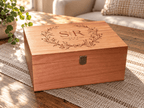 A rectangular wooden keepsake box with a hinged lid, featuring an engraved floral wreath and the initials 'S & R' with 'Special Pride' and '1994' on the lid. The box has a small metal latch on the front and is placed on a woven placemat on a wooden table in a sunlit living room setting.