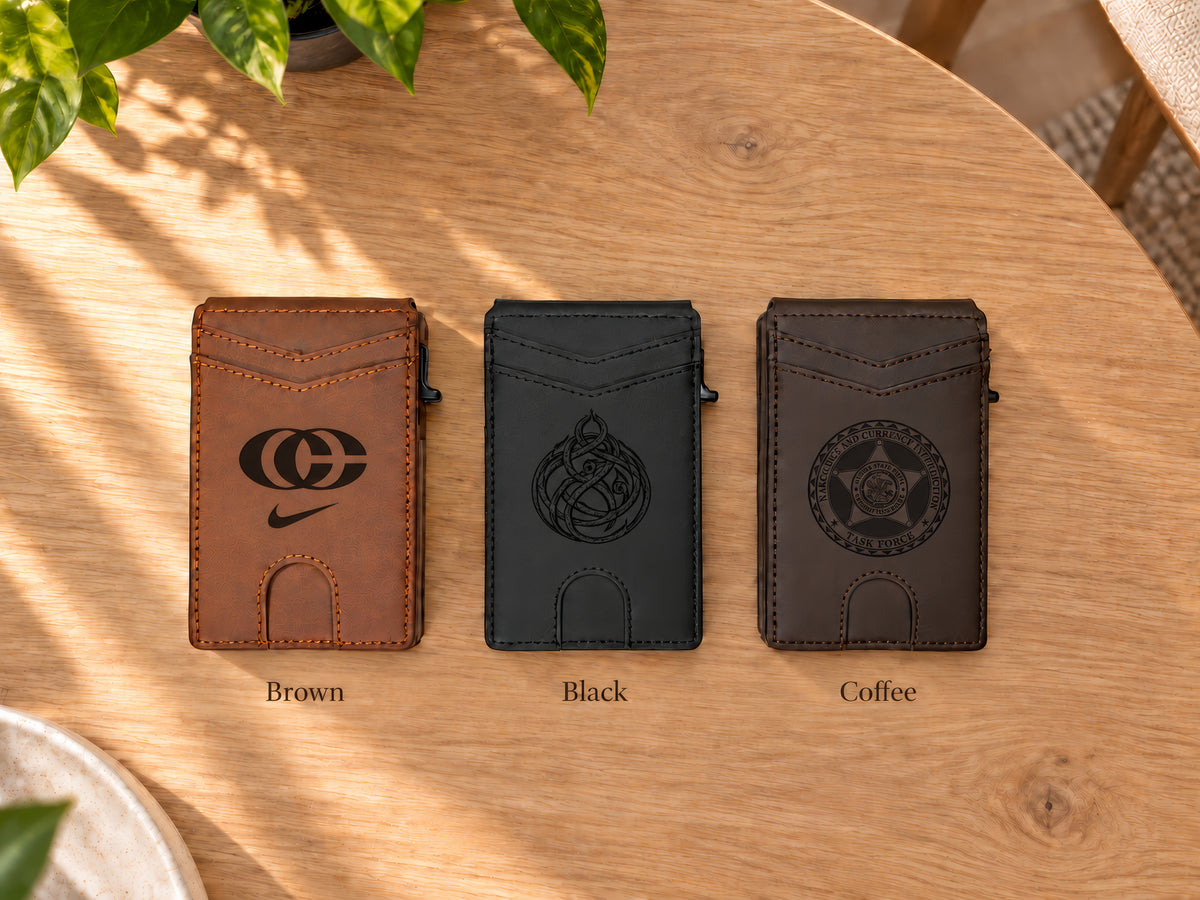 Three slim card wallets in brown, black, and dark coffee brown are lined up on a light wood table. Each wallet has an engraved logo on the front and a small leather loop at the bottom.